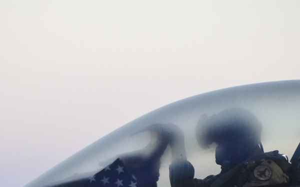A close-up view of a military fighter jet cockpit, likely an F-16 Fighting Falcon, showing one or two occupants wearing flight helmets and gear. An American flag is visible inside the cockpit. The image is taken at dusk or dawn, giving it a hazy, low-light quality.