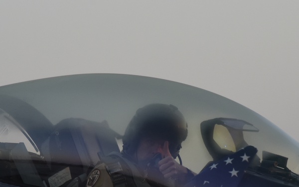 A military pilot is seated in the cockpit of a fighter jet, identifiable as an F-16 Fighting Falcon based on the canopy design and 'RESCUE' marking on the fuselage. The pilot is wearing a flight helmet and appears to be holding a folded American flag with visible white stars on a blue field. The image was taken from outside the aircraft at close range on an overcast day.