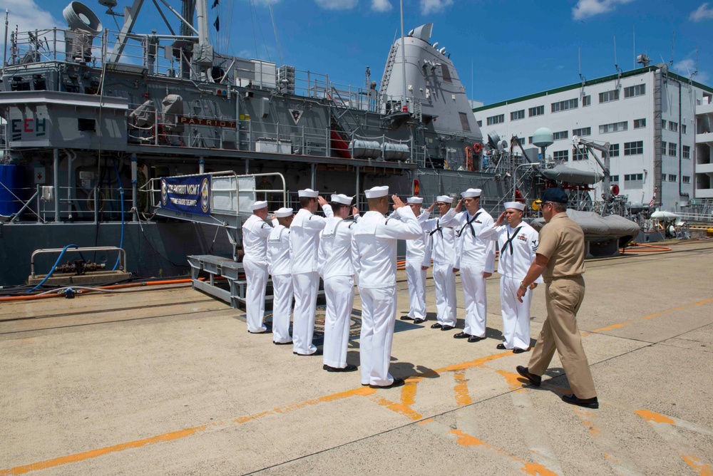 A formal naval ceremony is taking place on a pier alongside a warship, with a group of sailors in white dress uniforms rendering salutes to a senior officer in tan/khaki uniform. A banner on the ship reads 'USS PATRIOT MCM 7', identifying the vessel as a mine countermeasures ship. The setting appears to be a naval base, likely in Japan based on architectural elements visible in the background.