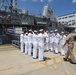 A formal naval ceremony is taking place on a pier alongside a warship, with a group of sailors in white dress uniforms rendering salutes to a senior officer in tan/khaki uniform. A banner on the ship reads 'USS PATRIOT MCM 7', identifying the vessel as a mine countermeasures ship. The setting appears to be a naval base, likely in Japan based on architectural elements visible in the background.