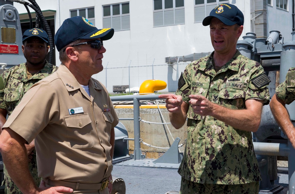 Three U.S. Navy personnel are gathered on what appears to be the deck of a naval vessel at a pier or naval installation. A sailor identified by name tape as 'HANSON' in camouflage NWU (Navy Working Uniform) is speaking and gesturing to a senior officer in khaki service uniform. A third sailor identified as 'KENNER' stands in the background.