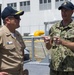 Three U.S. Navy personnel are gathered on what appears to be the deck of a naval vessel at a pier or naval installation. A sailor identified by name tape as 'HANSON' in camouflage NWU (Navy Working Uniform) is speaking and gesturing to a senior officer in khaki service uniform. A third sailor identified as 'KENNER' stands in the background.