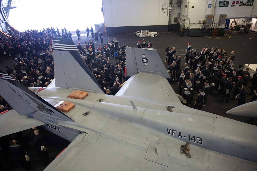 The image shows a large gathering of Navy personnel inside what appears to be an aircraft carrier hangar bay. Multiple F/A-18 Hornet jets belonging to Strike Fighter Squadron VFA-143 (the 'Pukin Dogs') are visible in the foreground, with hundreds of sailors assembled around them. The setting suggests a formal all-hands muster, briefing, or special event aboard the vessel.