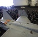 The image shows a large gathering of Navy personnel inside what appears to be an aircraft carrier hangar bay. Multiple F/A-18 Hornet jets belonging to Strike Fighter Squadron VFA-143 (the 'Pukin Dogs') are visible in the foreground, with hundreds of sailors assembled around them. The setting suggests a formal all-hands muster, briefing, or special event aboard the vessel.