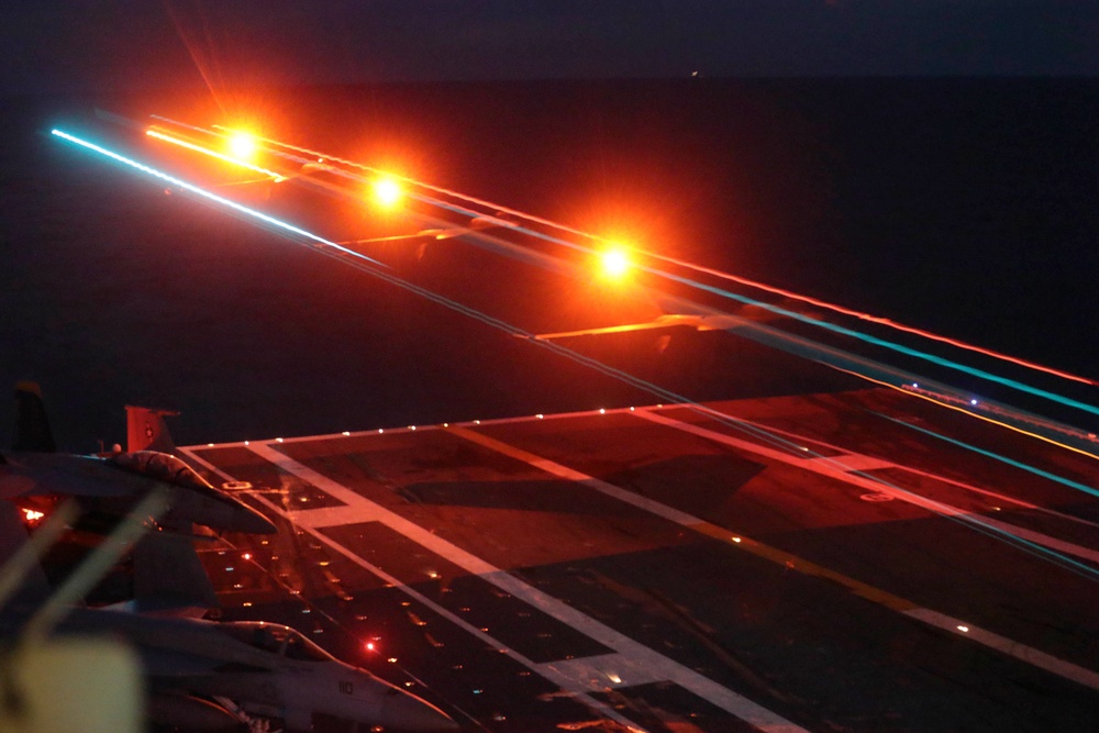 A long-exposure night photograph taken on what appears to be an aircraft carrier flight deck, capturing light trails from aircraft navigation and position lights during launch or landing operations. The image shows the characteristic markings and lighting of a carrier deck with streaking orange, red, and cyan/blue light trails indicating aircraft movement. The long shutter speed has created dramatic light streaks across the dark sky above the illuminated deck surface.