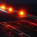 A long-exposure night photograph taken on what appears to be an aircraft carrier flight deck, capturing light trails from aircraft navigation and position lights during launch or landing operations. The image shows the characteristic markings and lighting of a carrier deck with streaking orange, red, and cyan/blue light trails indicating aircraft movement. The long shutter speed has created dramatic light streaks across the dark sky above the illuminated deck surface.