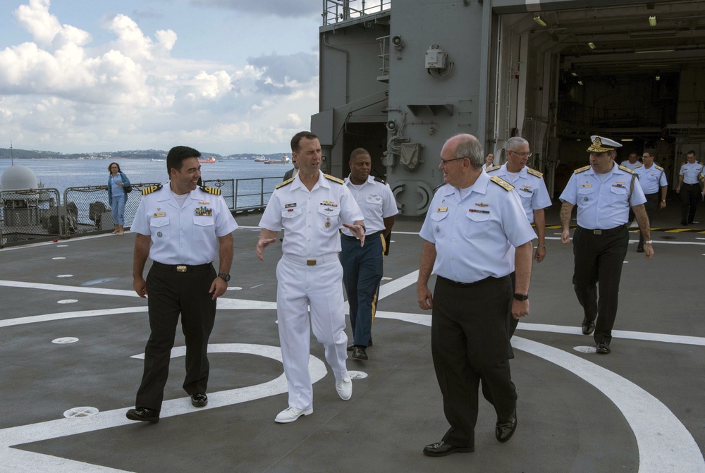 A group of naval officers in dress uniforms are walking on what appears to be a helicopter deck of a military vessel. The central figure is wearing full white dress uniform, while others wear white short-sleeve shirts with dark trousers, suggesting a formal visit or inspection. The setting appears to be a port with water and distant shoreline visible in the background.