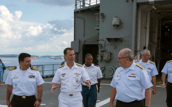 A group of naval officers in dress uniforms are walking on what appears to be a helicopter deck of a military vessel. The central figure is wearing full white dress uniform, while others wear white short-sleeve shirts with dark trousers, suggesting a formal visit or inspection. The setting appears to be a port with water and distant shoreline visible in the background.