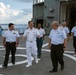 A group of naval officers in dress uniforms are walking on what appears to be a helicopter deck of a military vessel. The central figure is wearing full white dress uniform, while others wear white short-sleeve shirts with dark trousers, suggesting a formal visit or inspection. The setting appears to be a port with water and distant shoreline visible in the background.
