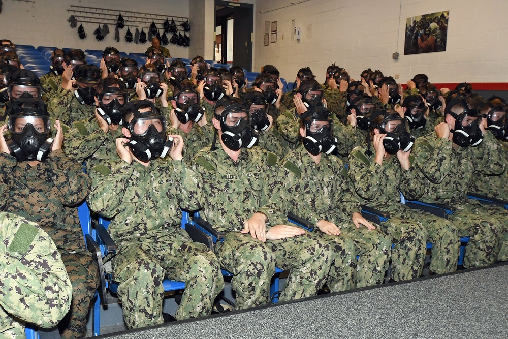 A large group of military personnel, primarily U.S. Navy sailors in NWU Type III (AOR-2) camouflage uniforms, are seated in an auditorium or training facility wearing black gas masks. The personnel appear to be participating in CBRN (Chemical, Biological, Radiological, and Nuclear) defense training, practicing donning and fitting their protective masks. At least one individual in the front left appears to be wearing USMC MARPAT digital camouflage.