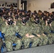 A large group of military personnel, primarily U.S. Navy sailors in NWU Type III (AOR-2) camouflage uniforms, are seated in an auditorium or training facility wearing black gas masks. The personnel appear to be participating in CBRN (Chemical, Biological, Radiological, and Nuclear) defense training, practicing donning and fitting their protective masks. At least one individual in the front left appears to be wearing USMC MARPAT digital camouflage.