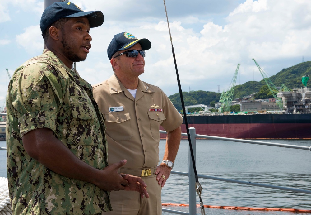 Two U.S. Navy personnel are standing on the deck of a vessel at a naval port facility, appearing to be in conversation. One is wearing Navy Working Uniform (NWU) camouflage and the other is in khaki service uniform with ribbons and a warfare device badge. The background shows a harbor environment with green cranes, a large ship hull, and tree-covered hills consistent with a naval base in Japan.