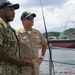 Two U.S. Navy personnel are standing on the deck of a vessel at a naval port facility, appearing to be in conversation. One is wearing Navy Working Uniform (NWU) camouflage and the other is in khaki service uniform with ribbons and a warfare device badge. The background shows a harbor environment with green cranes, a large ship hull, and tree-covered hills consistent with a naval base in Japan.