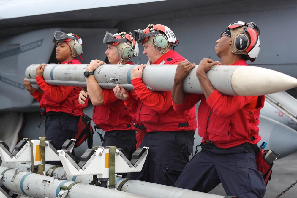 Four military personnel in red jerseys and flight deck helmets with hearing protection are carrying a missile or air-to-air weapon across what appears to be an aircraft carrier flight deck. A military jet aircraft, likely an F/A-18, is visible in the background. Additional ordnance on transport carts is visible in the foreground.