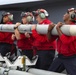 Four military personnel in red jerseys and flight deck helmets with hearing protection are carrying a missile or air-to-air weapon across what appears to be an aircraft carrier flight deck. A military jet aircraft, likely an F/A-18, is visible in the background. Additional ordnance on transport carts is visible in the foreground.