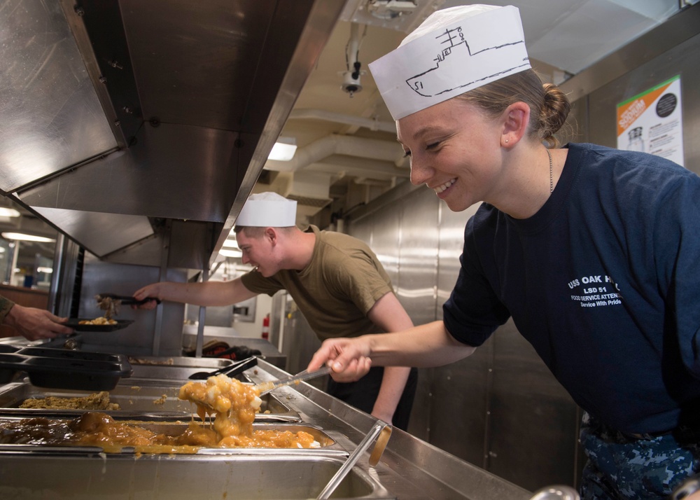 Two U.S. Navy personnel are serving food in a military ship galley (mess hall). The female sailor in the foreground is wearing a navy blue shirt identifying her as a Food Service Attendant aboard USS Oak Hill (LSD-51) and a paper sailor hat. Both individuals are actively serving food from steam table trays in a stainless steel kitchen environment.