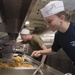 Two U.S. Navy personnel are serving food in a military ship galley (mess hall). The female sailor in the foreground is wearing a navy blue shirt identifying her as a Food Service Attendant aboard USS Oak Hill (LSD-51) and a paper sailor hat. Both individuals are actively serving food from steam table trays in a stainless steel kitchen environment.