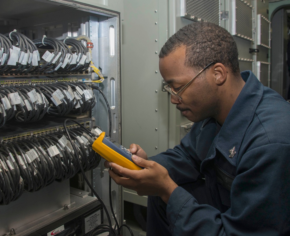 A technician in a dark blue uniform is using a yellow and gray handheld testing device on a network or communications equipment rack. The rack contains numerous bundled and labeled cables organized across multiple patch panel rows. The setting appears to be a technical or military facility based on the uniform insignia and equipment style.