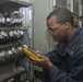 A technician in a dark blue uniform is using a yellow and gray handheld testing device on a network or communications equipment rack. The rack contains numerous bundled and labeled cables organized across multiple patch panel rows. The setting appears to be a technical or military facility based on the uniform insignia and equipment style.