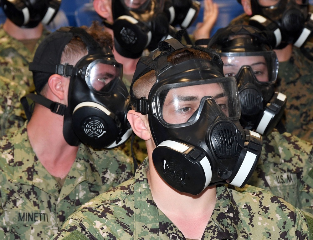 A group of U.S. Navy personnel wearing digital camouflage uniforms (NWU Type I) are fitted with black gas masks, likely during a CBRN (Chemical, Biological, Radiological, Nuclear) training exercise. Multiple sailors are seated in rows, all wearing identical protective respiratory equipment. A name tape reading 'MINETTI' and a partial 'U.S. NA[VY]' tape are visible on uniforms.