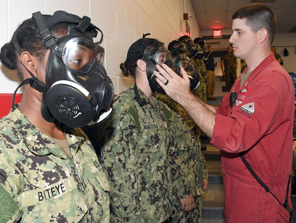 A military training exercise showing personnel in camouflage uniforms wearing gas masks being inspected by an instructor in a red jumpsuit. The scene appears to take place inside a military facility, with a line of personnel extending into the background. The instructor is checking the fit and seal of a gas mask on one of the trainees.
