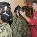 A military training exercise showing personnel in camouflage uniforms wearing gas masks being inspected by an instructor in a red jumpsuit. The scene appears to take place inside a military facility, with a line of personnel extending into the background. The instructor is checking the fit and seal of a gas mask on one of the trainees.
