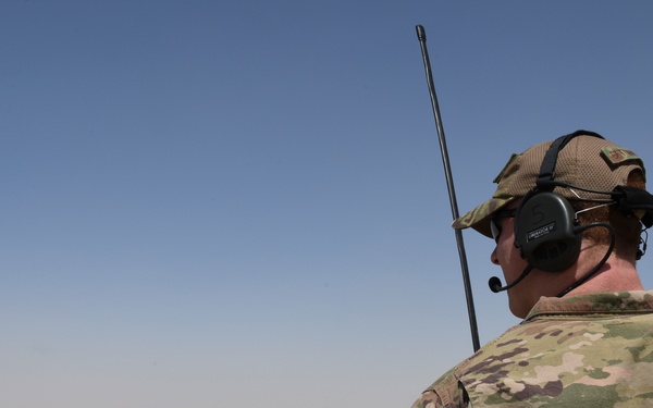 A military service member in MultiCam/OCP camouflage uniform is positioned on an elevated observation post overlooking a forward operating base in an arid desert environment. The soldier is equipped with tactical communications headset and a radio antenna, suggesting an active communications or overwatch role. The base below contains tents, sandbags, shipping containers, and a visible Red Cross medical marker.