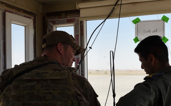Two military personnel are working inside what appears to be an airfield control tower or operations room, overlooking a tarmac area. One individual wears US Army multicam combat uniform with a patrol cap, while the other wears an olive/sage flight suit. A military transport aircraft (likely a C-130) is visible on the flight line through the windows.