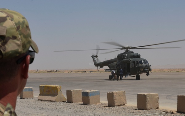 A U.S. Army soldier with the name tape 'SCHILL' visible on their cap observes a helicopter on a tarmac in what appears to be an arid, desert environment. The helicopter, identifiable as a Mi-17 or similar Soviet-designed transport helicopter, is on the ground with rotors spinning while ground crew personnel attend to it. Concrete barrier blocks line the perimeter separating the observation area from the flight line.