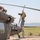 Three U.S. Army soldiers are performing maintenance or inspection on a military helicopter, identifiable by 'United States Army' markings on the fuselage. Two personnel are positioned on top of and near the tail rotor assembly, while a third stands on the ground near the tail boom. The setting appears to be a forward operating base or airfield in an arid environment.