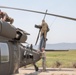 Three U.S. Army soldiers are performing maintenance or inspection on a military helicopter, identifiable by 'United States Army' markings on the fuselage. Two personnel are positioned on top of and near the tail rotor assembly, while a third stands on the ground near the tail boom. The setting appears to be a forward operating base or airfield in an arid environment.