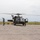 A military helicopter, consistent with a UH-60 Black Hawk, is on a concrete tarmac with its rotors spinning. At least two personnel in military uniforms are present — one appears to be signaling with raised arms to the left of the aircraft, and another is near the right side door. The setting appears to be a remote or forward operating airfield with flat terrain and mountains visible in the background.