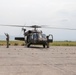 A military helicopter, consistent with a UH-60 Black Hawk, is on a concrete tarmac with its rotors spinning. At least two personnel in military uniforms are present — one appears to be signaling with raised arms to the left of the aircraft, and another is near the right side door. The setting appears to be a remote or forward operating airfield with flat terrain and mountains visible in the background.