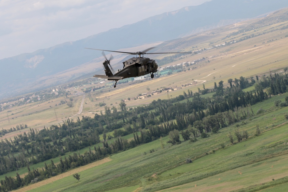 A dark military helicopter, consistent with a UH-60 Black Hawk design, is captured in flight over a green valley landscape. The terrain below features agricultural fields, tree lines, and a distant urban or semi-urban settlement backed by mountains. The image appears to be taken from another airborne platform or elevated position.