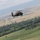 A dark military helicopter, consistent with a UH-60 Black Hawk design, is captured in flight over a green valley landscape. The terrain below features agricultural fields, tree lines, and a distant urban or semi-urban settlement backed by mountains. The image appears to be taken from another airborne platform or elevated position.