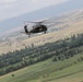 A dark military helicopter, consistent with a UH-60 Black Hawk design, is captured in flight over a green valley landscape. The terrain below features agricultural fields, tree lines, and a distant urban or semi-urban settlement backed by mountains. The image appears to be taken from another airborne platform or elevated position.
