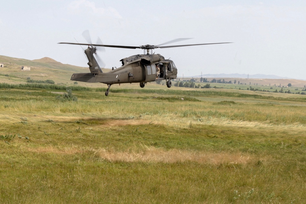 A military utility helicopter, consistent with a UH-60 Black Hawk, is hovering at low altitude over an open grassy field. The aircraft is painted in olive drab/dark green military livery and has its side door open with at least one crew member visible. The surrounding terrain appears to be rolling hills with sparse vegetation, suggesting a training or operational exercise environment.
