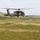 A military utility helicopter, consistent with a UH-60 Black Hawk, is hovering at low altitude over an open grassy field. The aircraft is painted in olive drab/dark green military livery and has its side door open with at least one crew member visible. The surrounding terrain appears to be rolling hills with sparse vegetation, suggesting a training or operational exercise environment.