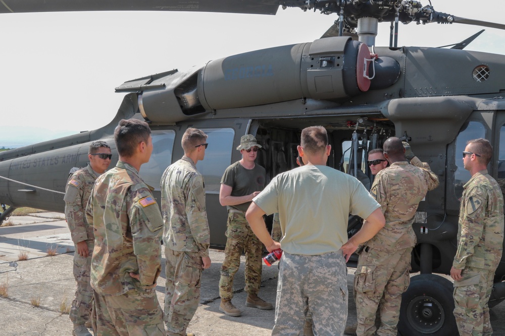 A group of approximately nine U.S. Army personnel are gathered around a UH-60 Black Hawk helicopter on a tarmac or airfield. One individual in a bucket hat and olive t-shirt appears to be briefing or instructing the group near the open cabin door of the aircraft. The helicopter bears 'UNITED STATES ARMY' markings and is painted in standard olive drab camouflage.