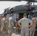 A group of approximately nine U.S. Army personnel are gathered around a UH-60 Black Hawk helicopter on a tarmac or airfield. One individual in a bucket hat and olive t-shirt appears to be briefing or instructing the group near the open cabin door of the aircraft. The helicopter bears 'UNITED STATES ARMY' markings and is painted in standard olive drab camouflage.