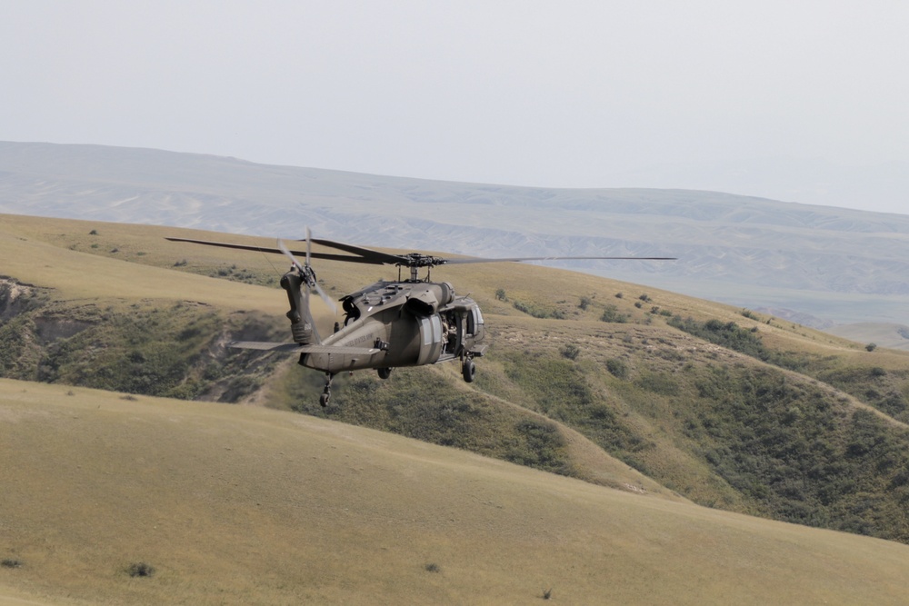 A military helicopter, consistent with a UH-60 Black Hawk, is flying low over rolling, semi-arid hills with sparse vegetation. The aircraft is in an olive drab/gray military paint scheme and appears to be on a low-altitude flight mission. The background features layered terrain with distant ridgelines under a hazy sky.