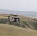 A military helicopter, consistent with a UH-60 Black Hawk, is flying low over rolling, semi-arid hills with sparse vegetation. The aircraft is in an olive drab/gray military paint scheme and appears to be on a low-altitude flight mission. The background features layered terrain with distant ridgelines under a hazy sky.