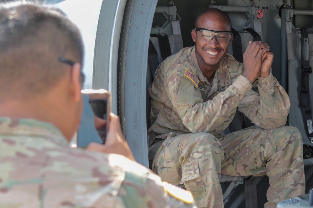 A U.S. Army soldier in OCP (Operational Camouflage Pattern) uniform sits smiling inside the open door of a military helicopter or aircraft, wearing clear safety glasses. Another soldier in the foreground is photographing him with a smartphone. The scene appears informal and lighthearted, likely during a training exercise or airfield visit.