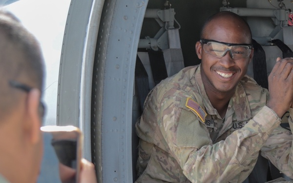 A U.S. Army soldier in OCP (Operational Camouflage Pattern) uniform sits smiling inside the open door of a military helicopter or aircraft, wearing clear safety glasses. Another soldier in the foreground is photographing him with a smartphone. The scene appears informal and lighthearted, likely during a training exercise or airfield visit.