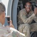 A U.S. Army soldier in OCP (Operational Camouflage Pattern) uniform sits smiling inside the open door of a military helicopter or aircraft, wearing clear safety glasses. Another soldier in the foreground is photographing him with a smartphone. The scene appears informal and lighthearted, likely during a training exercise or airfield visit.