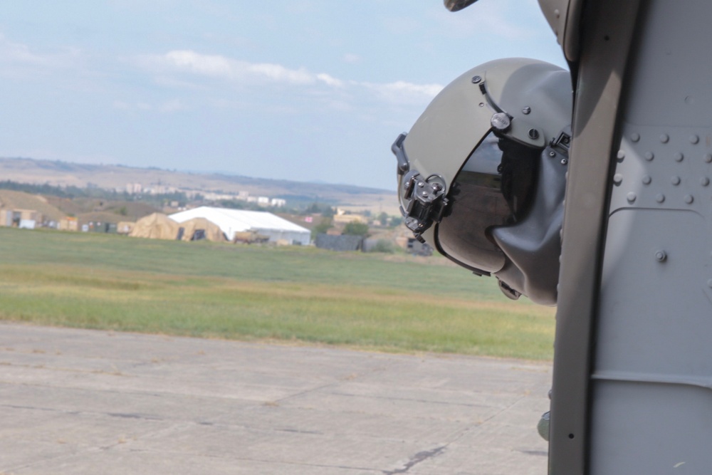 The image shows a military flight helmet mounted or resting against the exterior fuselage of a military helicopter on an airfield. The helmet appears to be equipped with night vision goggle (NVG) mounts and a dark visor. In the background, a military airfield is visible with tents, structures, and open grassy terrain.