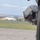 The image shows a military flight helmet mounted or resting against the exterior fuselage of a military helicopter on an airfield. The helmet appears to be equipped with night vision goggle (NVG) mounts and a dark visor. In the background, a military airfield is visible with tents, structures, and open grassy terrain.