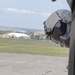 The image shows a military flight helmet mounted or resting against the exterior fuselage of a military helicopter on an airfield. The helmet appears to be equipped with night vision goggle (NVG) mounts and a dark visor. In the background, a military airfield is visible with tents, structures, and open grassy terrain.