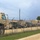 A large number of military vehicles, primarily tan/desert-colored tactical trucks and HMMWVs (Humvees), are parked in a secured motor pool behind a chain-link fence topped with barbed wire. The vehicles appear to be staged in organized rows on a gravel/dirt surface, consistent with a military installation or National Guard armory. The photo is taken from outside the perimeter fence along a dirt road.