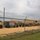 A secured military vehicle storage yard viewed from outside a chain-link fence topped with barbed wire. Multiple tan/olive drab military vehicles including HMMWVs (Humvees) and large cargo trucks are parked on a gravel/dirt surface. A warning sign on the fence reads 'WARNING RESTRICTED AREA - KEEP OUT - AUTHORIZED PERSONNEL ONLY'.